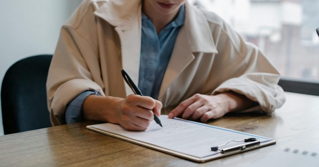 A woman in a formal setting fills out paperwork on a clipboard at an office desk.